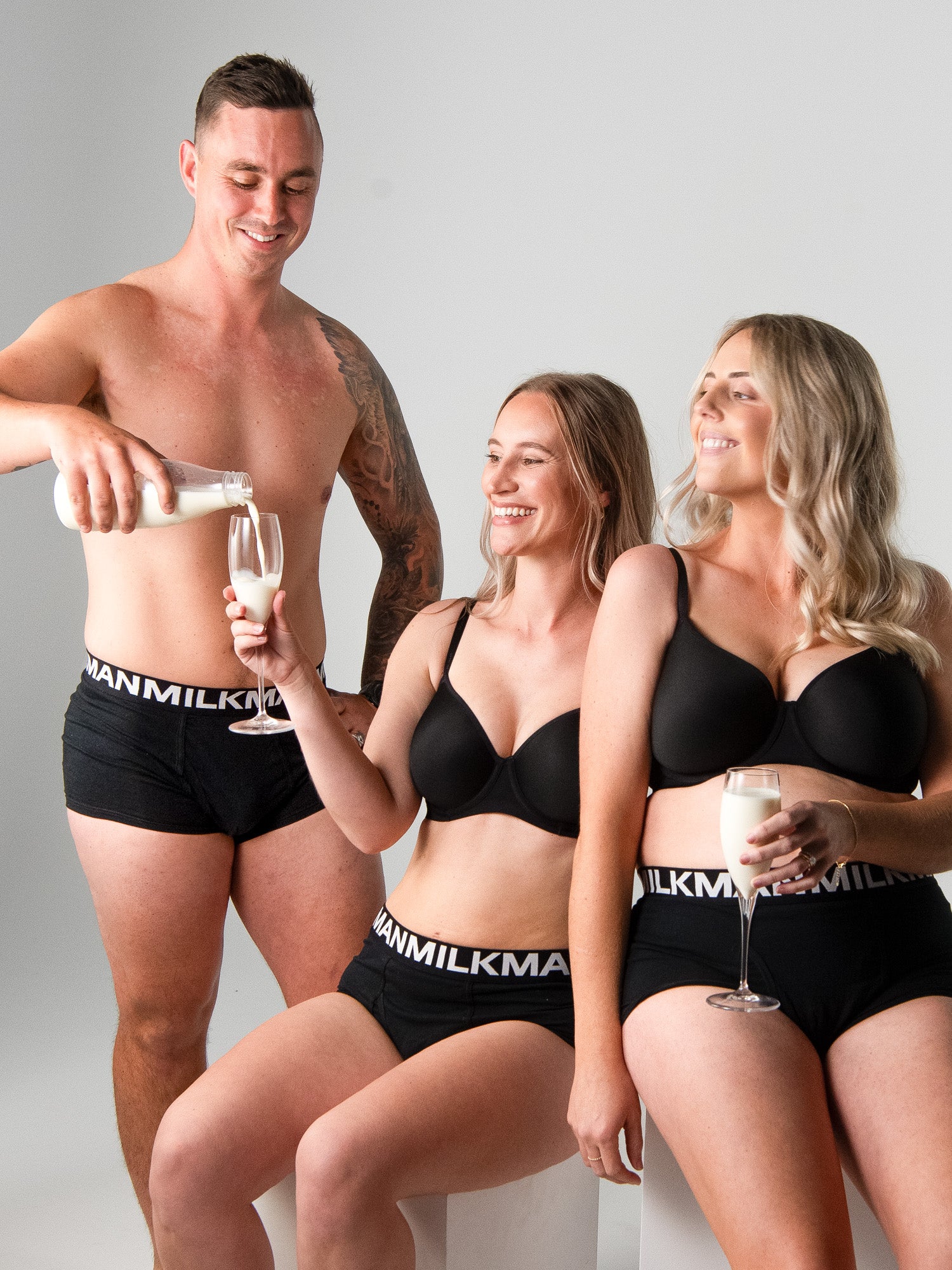 Two women in black underwear with 'Milkman' branding, one pouring milk into a glass held by a man.