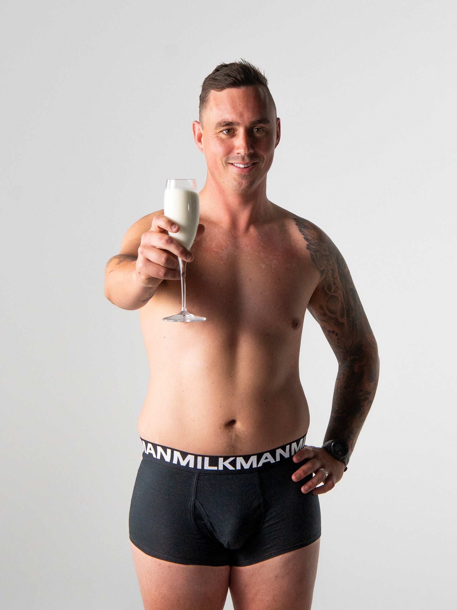 Man wearing black underwear with 'Milkman' branding, holding a glass of milk against a plain background.